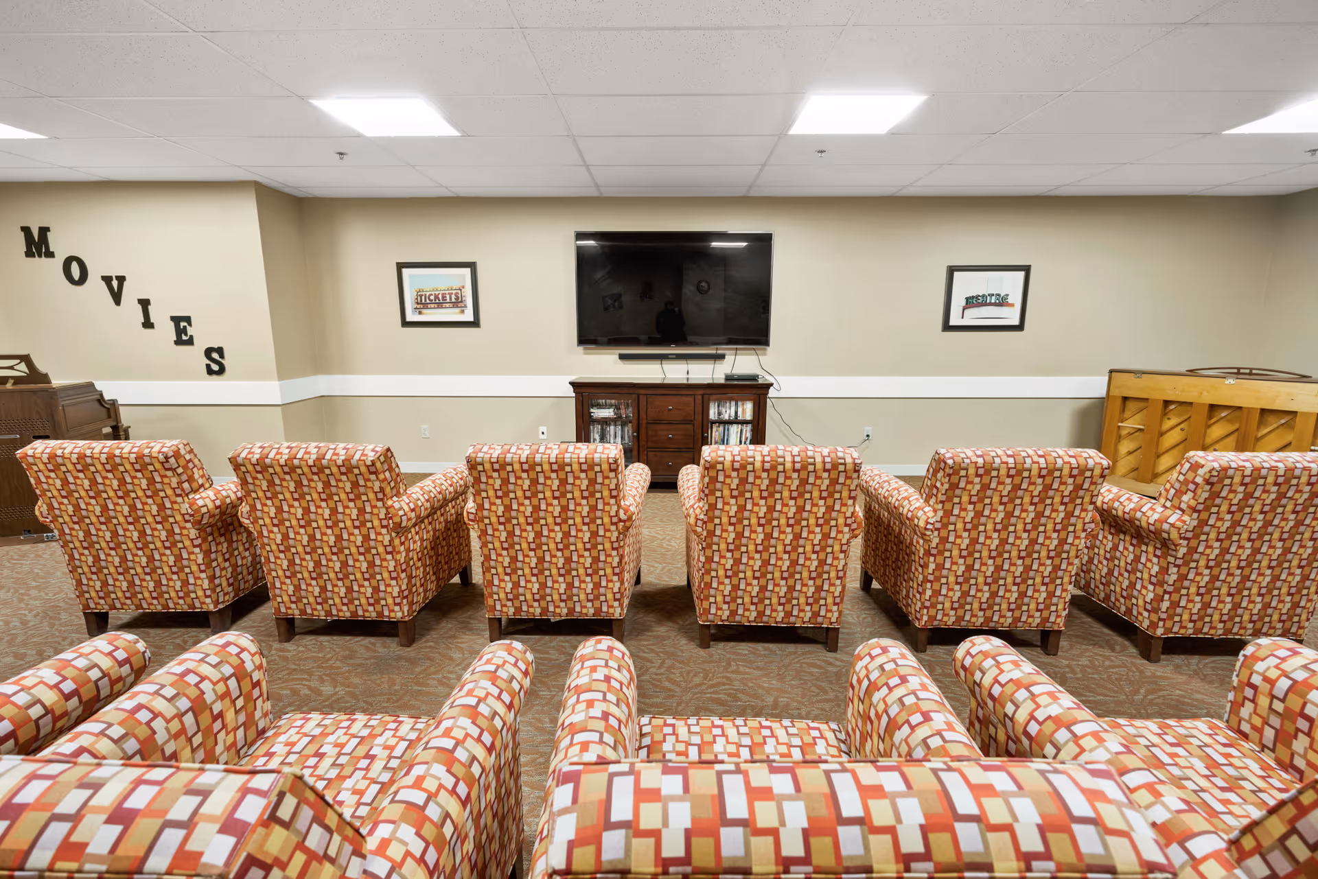 A small movie viewing room with two rows of patterned armchairs facing a flat-screen TV mounted on a beige wall. Below the TV is a wooden cabinet with DVDs. The word 'MOVIES' is displayed on the left wall, and there is a piano on the right side of the room.