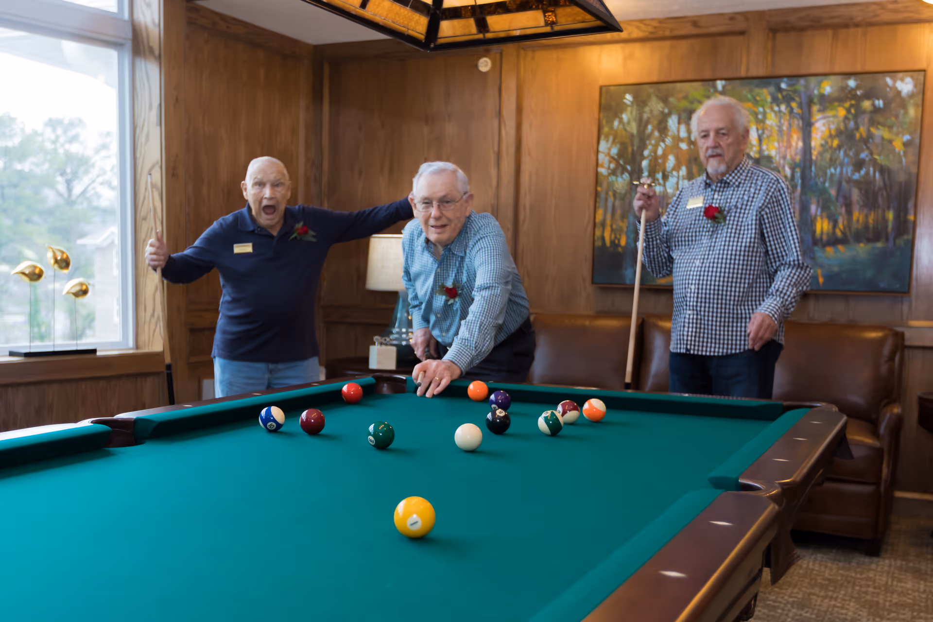 Three elderly men playing pool in a wood-paneled room with a large window. One man is taking a shot while the other two watch, one with a surprised expression and the other holding a pool cue. There is a painting of a forest on the wall and a brown leather couch in the background.