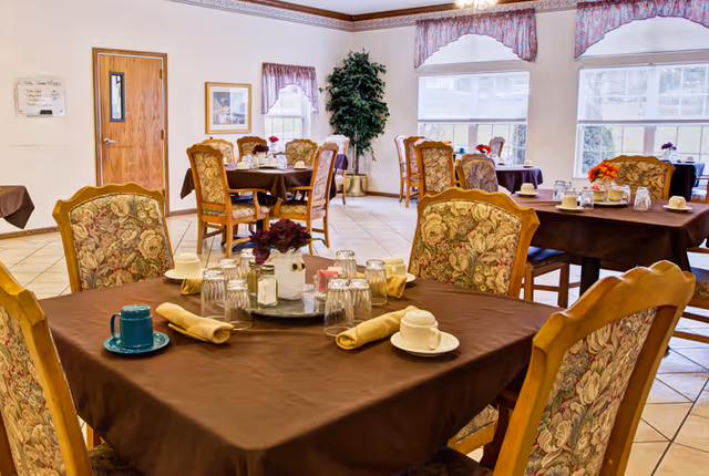 Bright dining room with several tables covered in brown tablecloths, floral-upholstered chairs, place settings, and large windows.