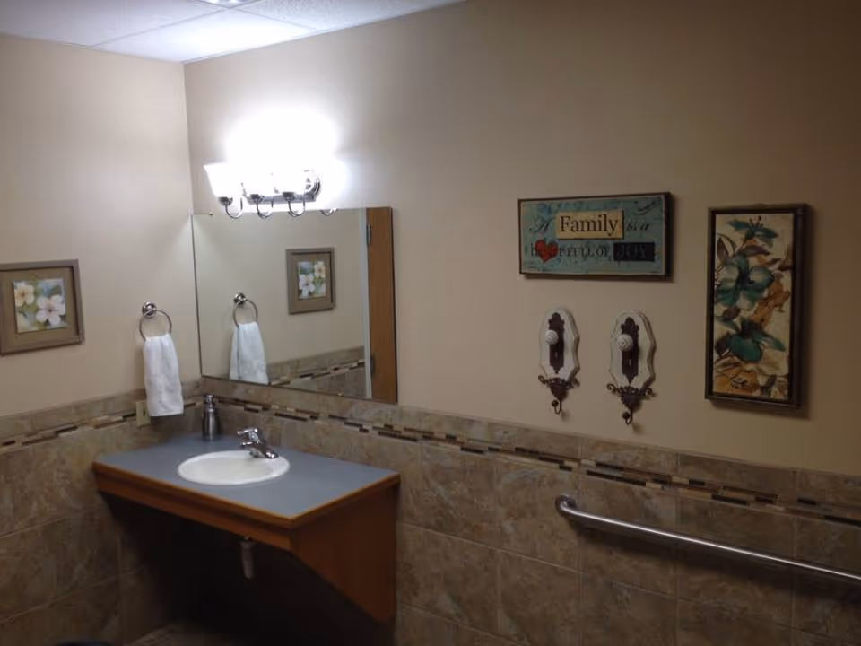 A bathroom interior with a wall-mounted sink and faucet, a large mirror above the sink, a towel ring holding a white towel, and decorative wall art including floral pictures and a sign that reads 'Family full of joy'. The walls are beige with a tiled lower half and a metal grab bar is mounted on the wall.