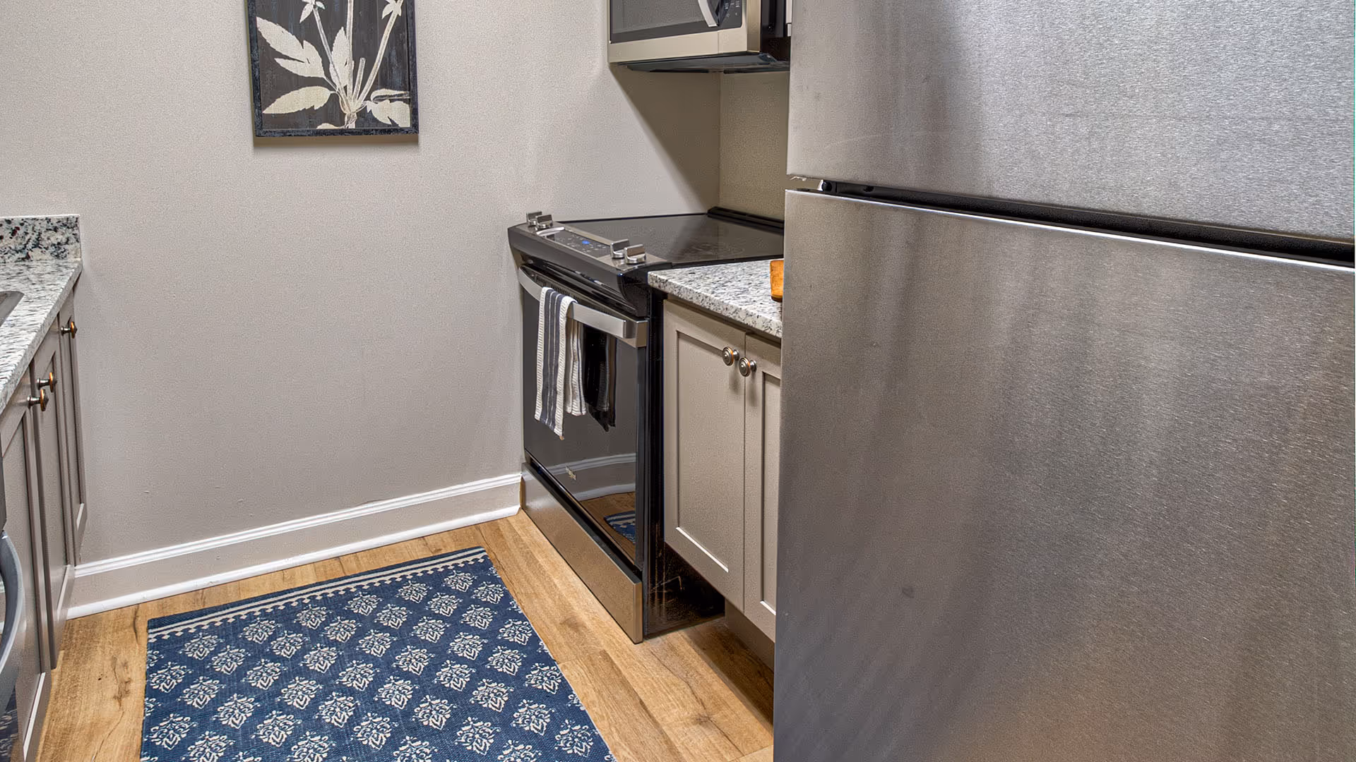 A modern kitchen with stainless steel appliances including a refrigerator and an electric stove. The kitchen features granite countertops, light-colored cabinets, a blue patterned rug on a wooden floor, and a framed botanical print on the wall.