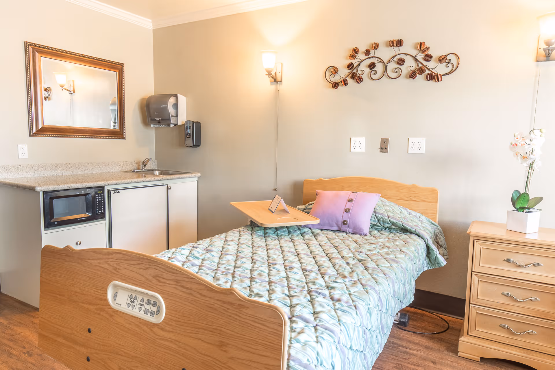 A senior living facility room with a single bed covered in a quilted blanket and a purple pillow. There is a wooden bedside table with a white orchid plant on it. On the left side, there is a small kitchenette area with a microwave, mini fridge, sink, and a mirror above the counter. The walls are light-colored with decorative wall art and two wall-mounted light fixtures.