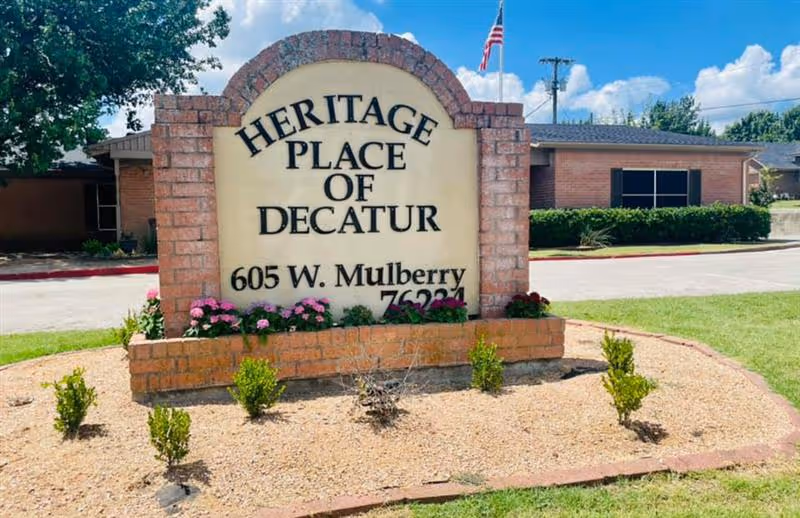Brick monument sign reading "Heritage Place of Decatur, 605 W. Mulberry" in front of a single-story building and an American flag.