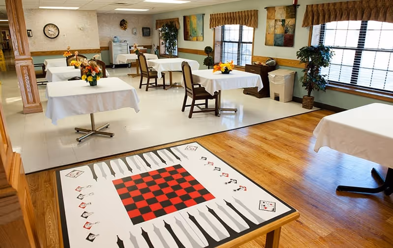 A bright dining room with several tables covered in white tablecloths, each adorned with a small floral centerpiece. The room has large windows with brown valances, wooden flooring, and a game table in the foreground featuring a checkerboard and backgammon design. The walls are decorated with paintings and a clock.