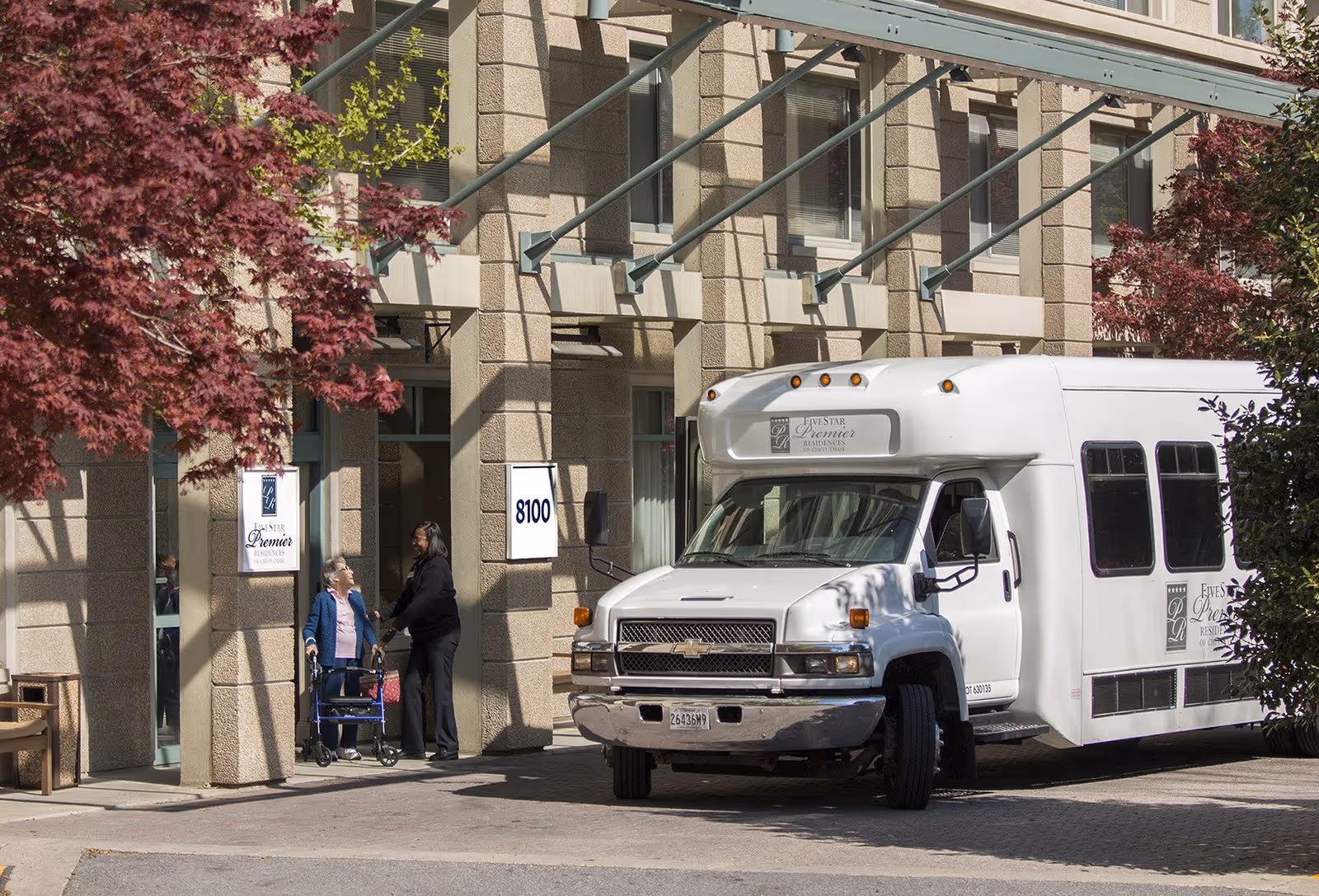 An elderly woman with a walker is talking to a staff member outside the entrance of Five Star Premier Residences of Chevy Chase. A white shuttle bus with the facility's logo is parked nearby. The building exterior features beige stone walls and large windows, with red and green trees framing the scene.