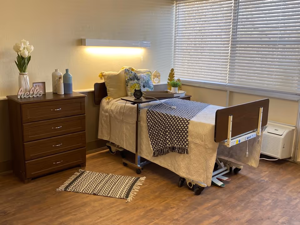 A neatly arranged room with a hospital-style bed covered with a white quilt and a black and white patterned throw. The bed has several pillows, including a floral one, and a small overbed table with a clock on it. Next to the bed is a wooden dresser with decorative items including a vase with white tulips, two ceramic bottles, a small framed picture, and a 'hello' sign. The room has wooden flooring, a small patterned rug, and large windows with blinds partially closed. A wall-mounted light fixture illuminates the bed area.