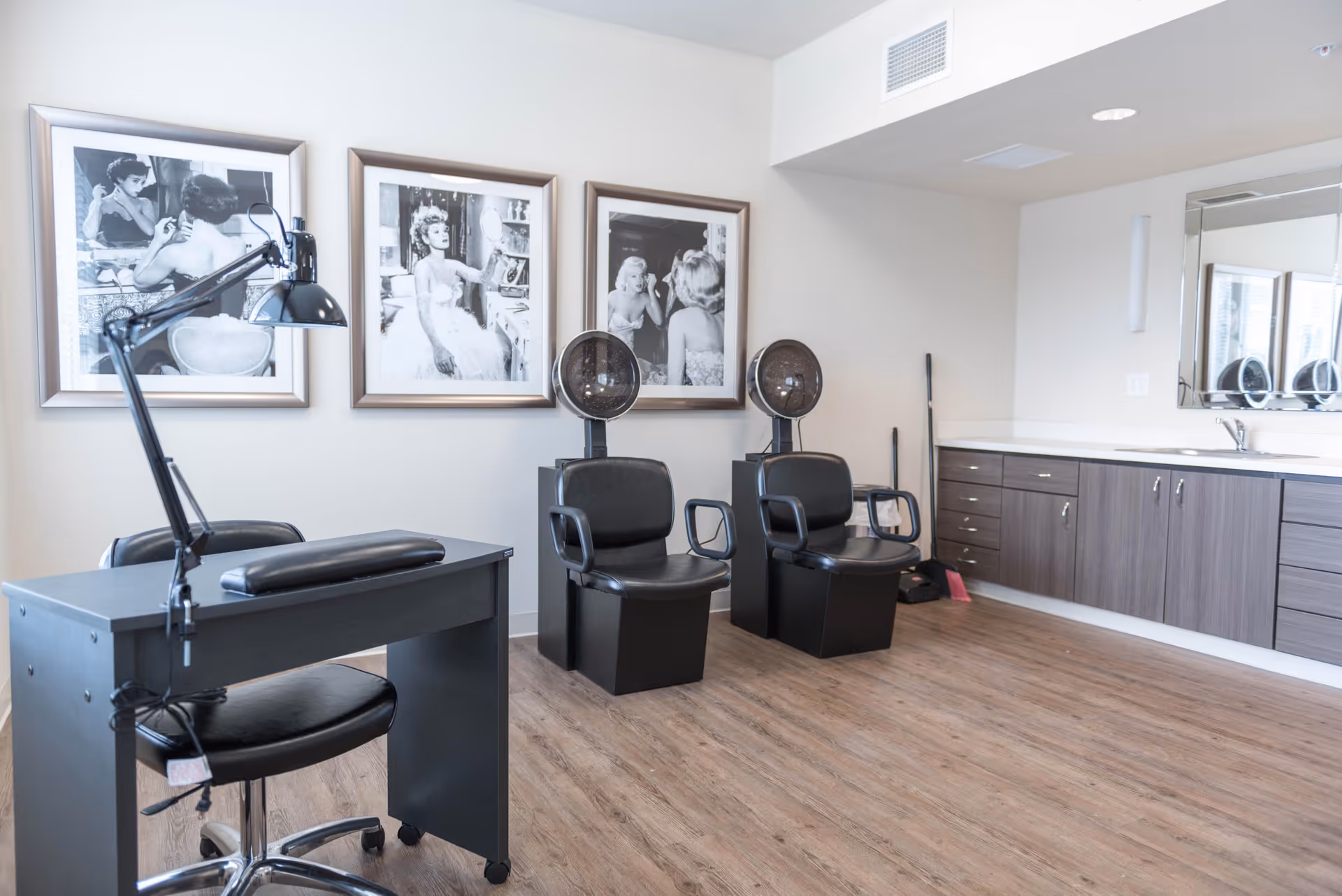 Interior of a salon area with two black hair drying chairs, a manicure table with a black lamp, and three black and white framed photos of women on the wall. There is a countertop with cabinets and a sink on the right side of the room.