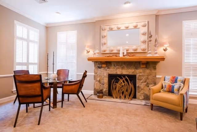 A cozy living room area with a stone fireplace featuring a decorative metal screen. Above the fireplace is a large mirror with a unique frame. To the left, there is a round glass table with four brown leather chairs. To the right, there is a beige armchair with a colorful pillow. The room has beige walls and carpet, with two windows featuring white plantation shutters.