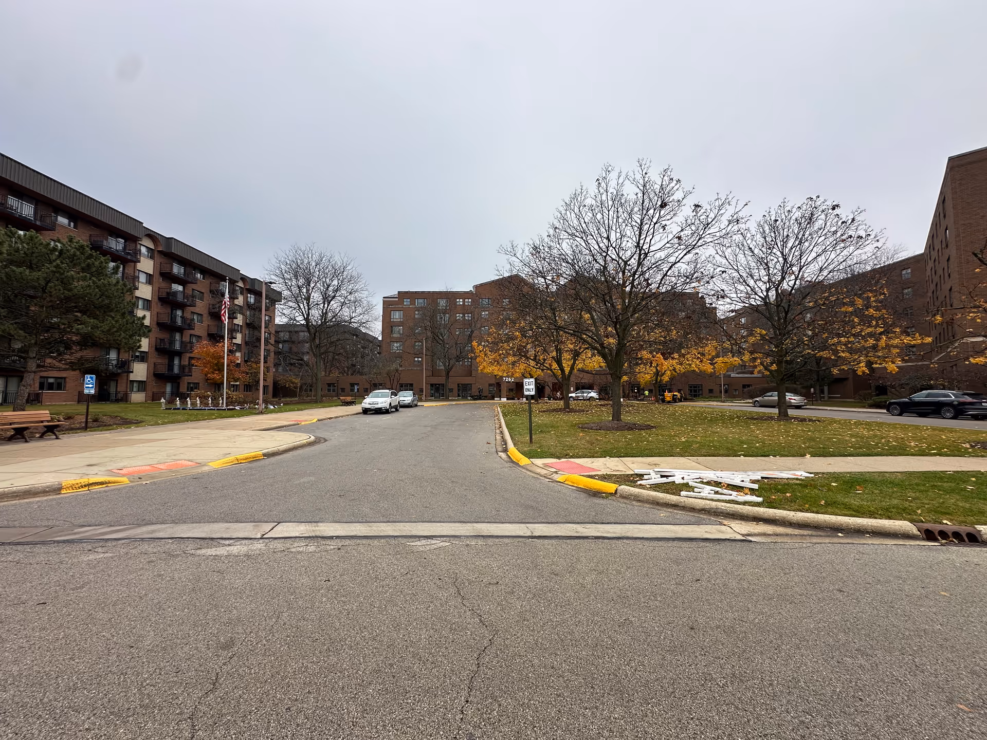 Front driveway and entrance of a multi-story brick senior living complex with trees, parked cars, and a cloudy sky.