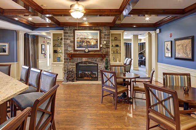 A cozy senior living common area with wooden floors, a stone fireplace with a mantel, and a framed painting above it. There are several wooden chairs with cushions around small round tables and a counter with high chairs. The walls are painted blue with white wainscoting, and the ceiling has wooden beams with a ceiling fan and lights.