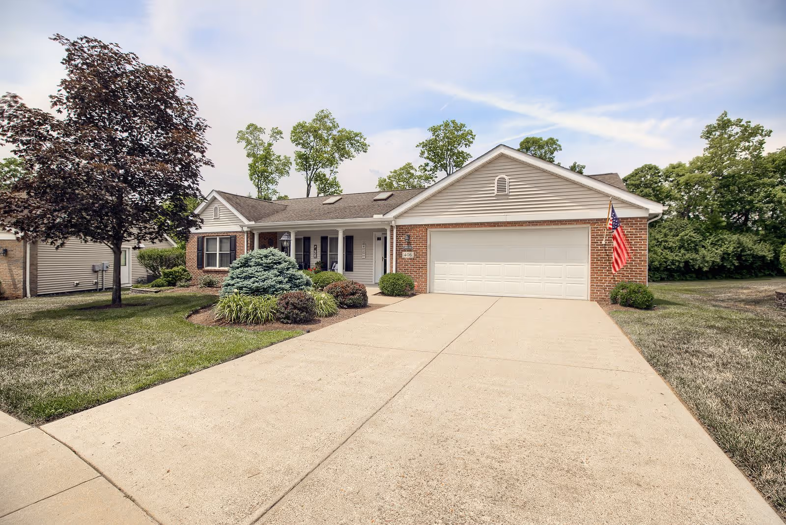Single-story residential building with a two-car garage, a concrete driveway, and a landscaped front yard featuring bushes and a tree. An American flag is mounted near the garage door. The sky is partly cloudy and there are trees in the background.