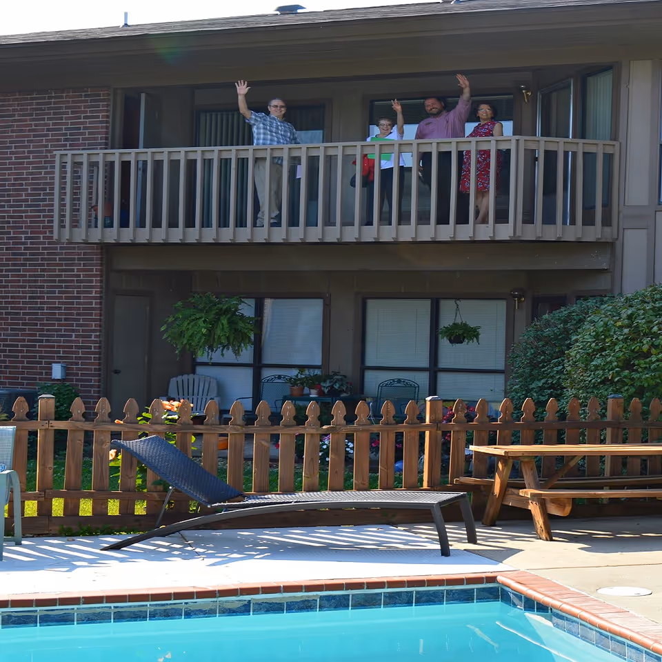 Four people standing on a second-floor balcony of a senior living facility waving. Below the balcony is a fenced garden area with plants, a lounge chair, a picnic table, and a swimming pool in the foreground.