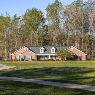 A single-story brick building with a gray roof and three dormer windows, surrounded by a large grassy lawn and trees in the background under a clear sky.