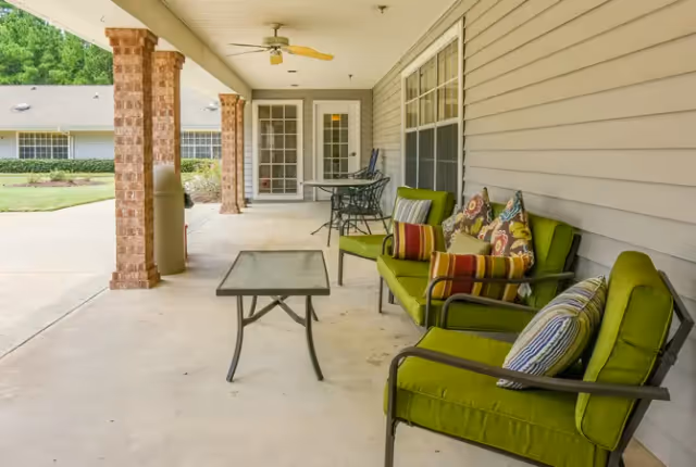 Covered porch with green cushioned chairs, a glass-top coffee table, and a small dining set overlooking a landscaped courtyard.