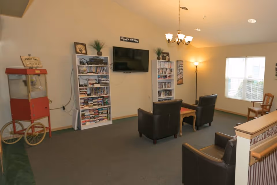 A senior living facility common area with a wall-mounted flat screen TV flanked by two white bookshelves filled with books and DVDs. There are three dark leather armchairs arranged around a wooden coffee table. A red popcorn machine is positioned on the left side of the room. The room is softly lit by a chandelier and a floor lamp, with a window letting in natural light on the right side.
