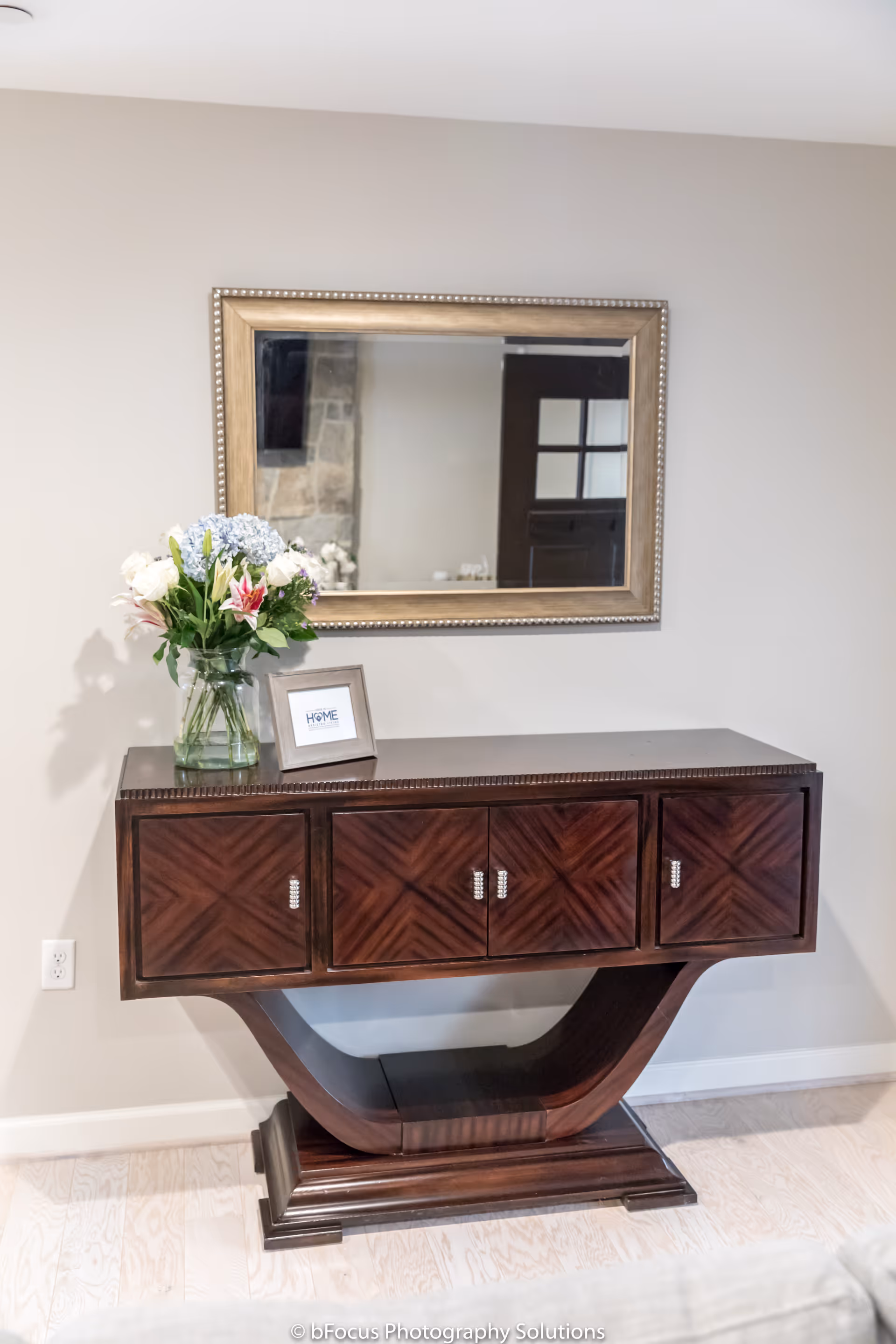A dark wooden console table with a curved base stands against a light-colored wall. On top of the table is a glass vase with a bouquet of flowers and a small framed sign that says 'HOME'. Above the table hangs a large rectangular mirror with a decorative frame. The floor is light wood, and a reflection in the mirror shows part of a stone wall and a door with glass panes.
