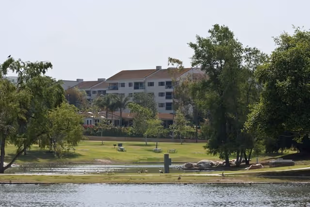 View of a senior living facility named Regents Point with a large multi-story building in the background surrounded by trees and greenery. In the foreground, there is a pond with a grassy area featuring picnic tables and benches.