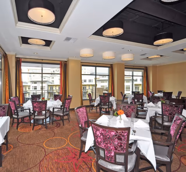 Dining room with multiple tables covered in white tablecloths, each set with napkins and glassware. The chairs have dark wood frames with purple patterned upholstery. Large windows with orange and red curtains allow natural light to fill the room. The ceiling features recessed lighting with circular fixtures.
