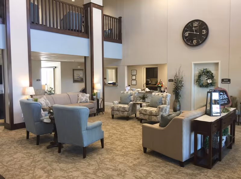 A spacious and well-lit living room area in an assisted living facility featuring multiple upholstered chairs and sofas arranged around small tables. The room has a high ceiling with a large wall clock, decorative plants, and a console table with a small sign and lantern. The decor is neutral and inviting, with carpeted flooring and wooden accents.