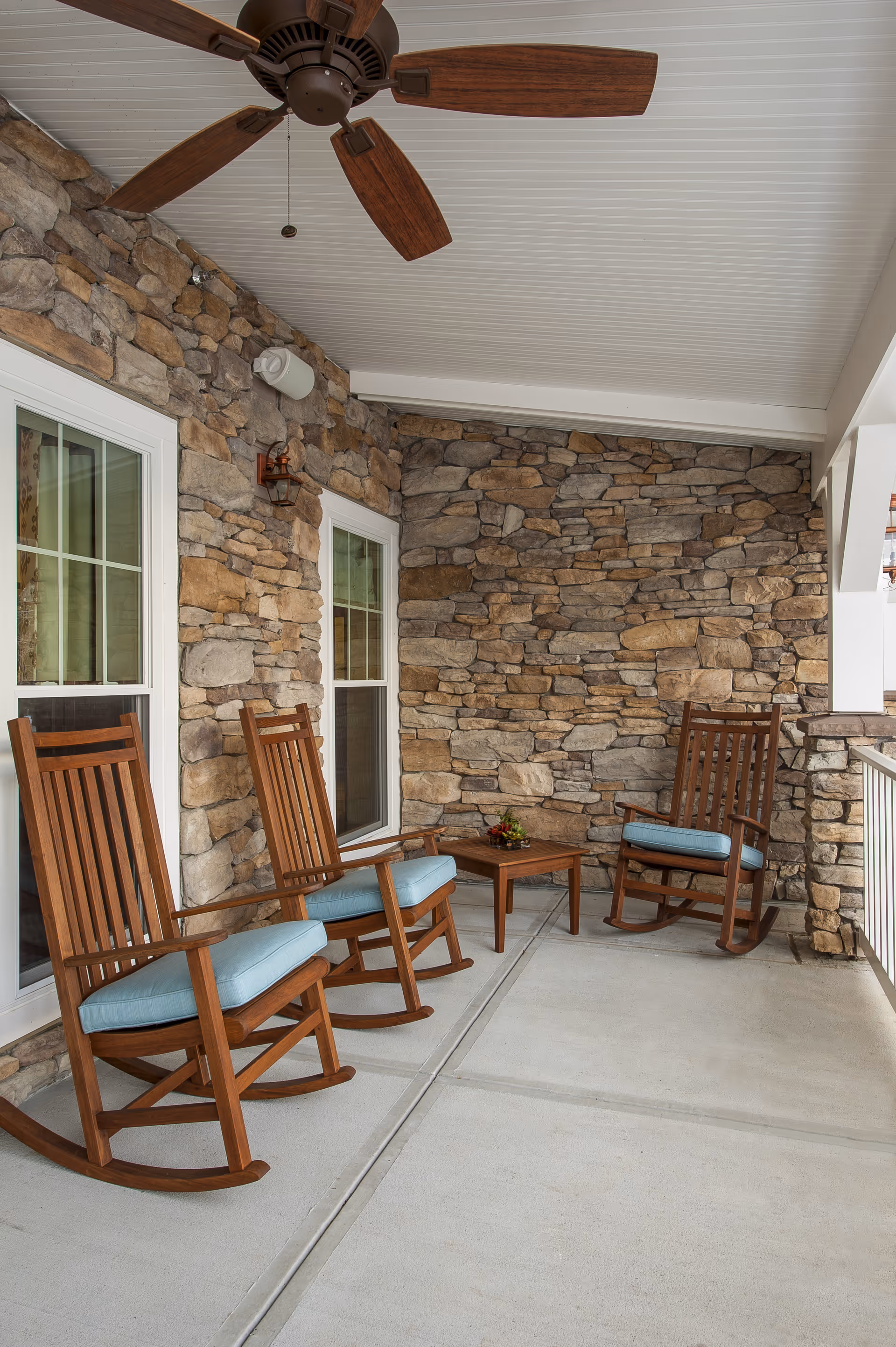 Covered outdoor patio area with stone walls, three wooden rocking chairs with blue cushions, a small wooden table with a flower arrangement, and a ceiling fan above.