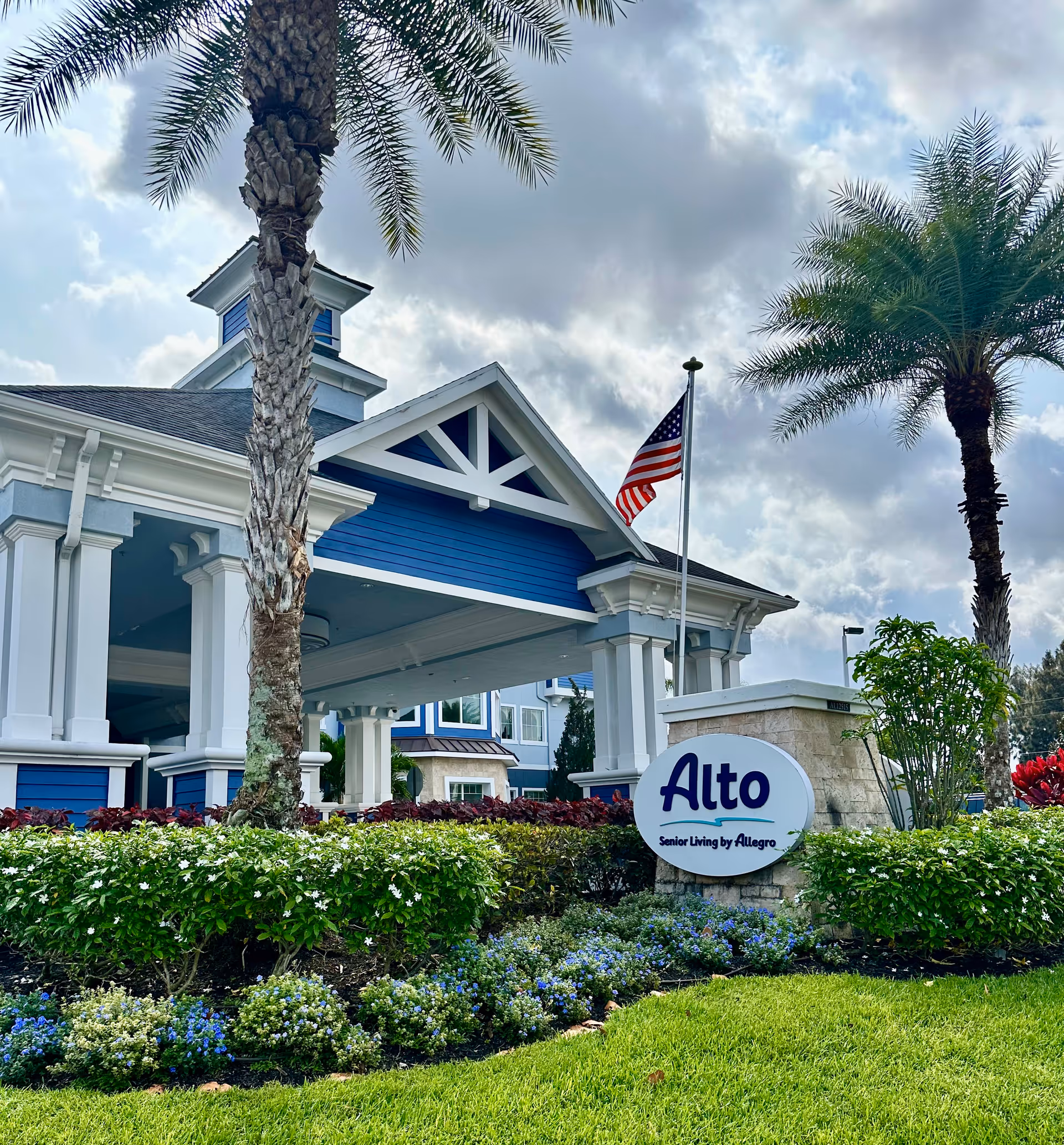 Exterior view of the Alto Stuart senior living facility entrance with a blue and white building, palm trees, an American flag, and a landscaped garden with green shrubs and colorful flowers.