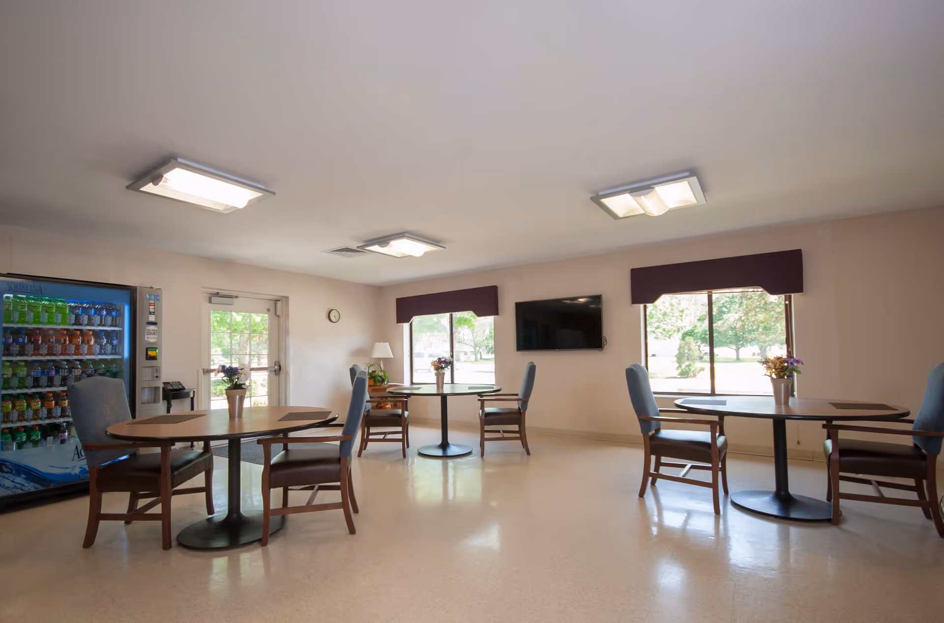 A bright and clean common dining area with three round tables, each surrounded by four wooden chairs with blue cushions. There are two large windows with purple valances, a wall-mounted flat-screen TV, and a vending machine stocked with beverages. A door with a window leads outside, and the floor is a light-colored linoleum.
