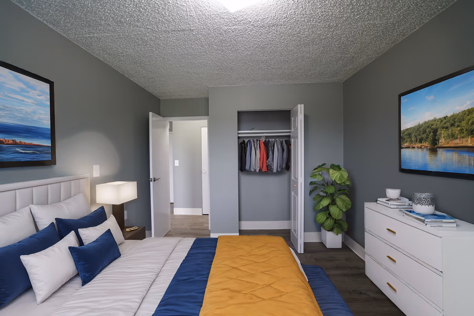 A neatly arranged bedroom with a bed featuring white and blue bedding with a yellow throw blanket. There is a nightstand with a lamp next to the bed, an open closet with hanging clothes, a white dresser with decorative items on top, a green potted plant, and two framed landscape paintings on the walls. The walls are painted gray and the floor is wooden.