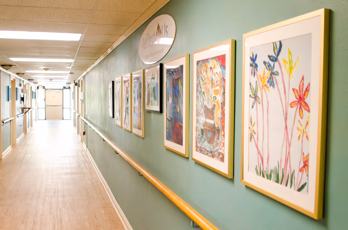 A brightly lit hallway in a healthcare facility with light wooden flooring and green walls. The right wall is decorated with framed colorful artwork and a sign that reads 'Reflect Nails'. Handrails run along both sides of the hallway, and there are doors and windows along the left side. The hallway leads to a set of double doors at the far end.