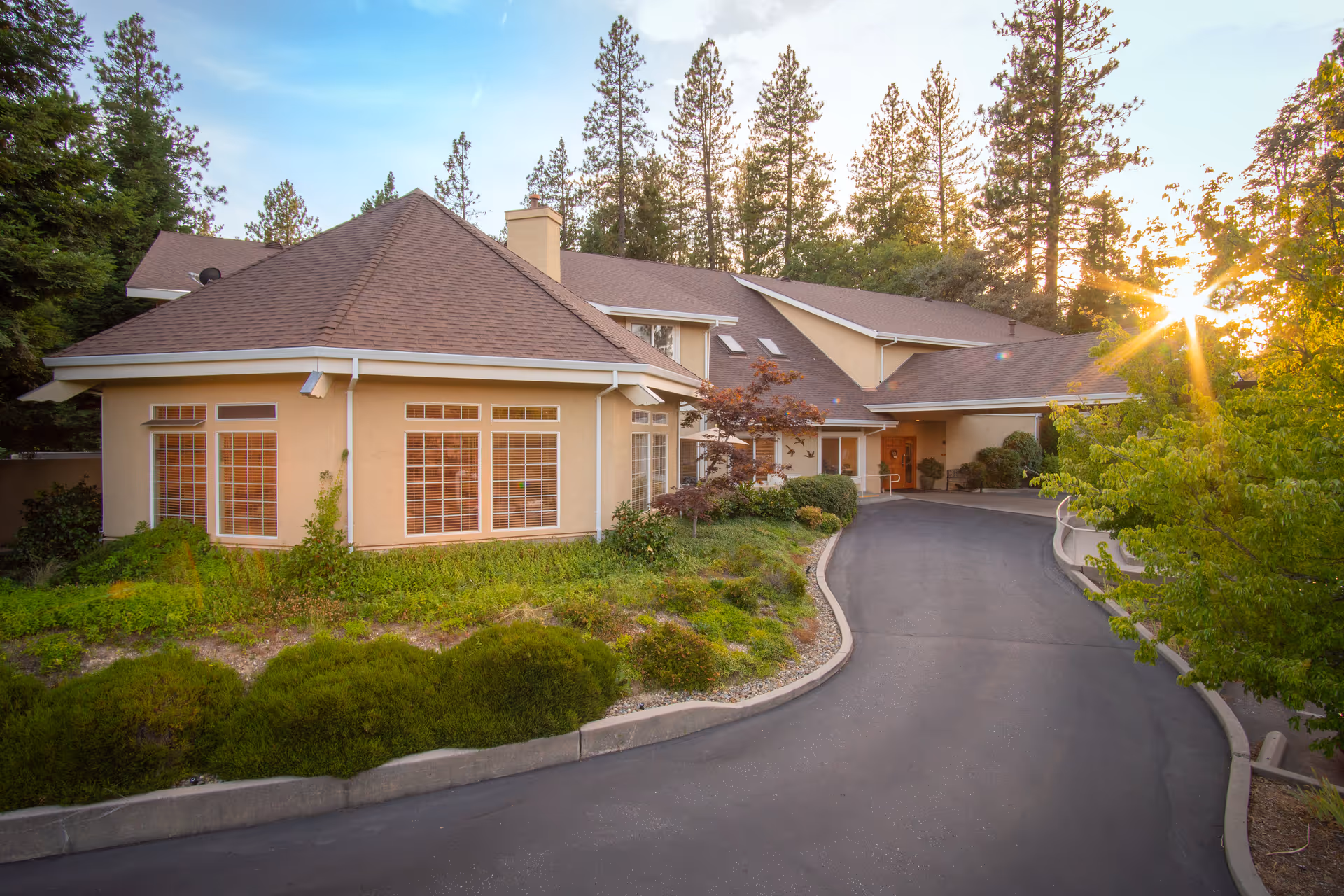 Exterior view of Brunswick Village Assisted Living facility at sunset, showing a beige building with large windows, surrounded by greenery and tall pine trees, with a curved driveway leading to the entrance.