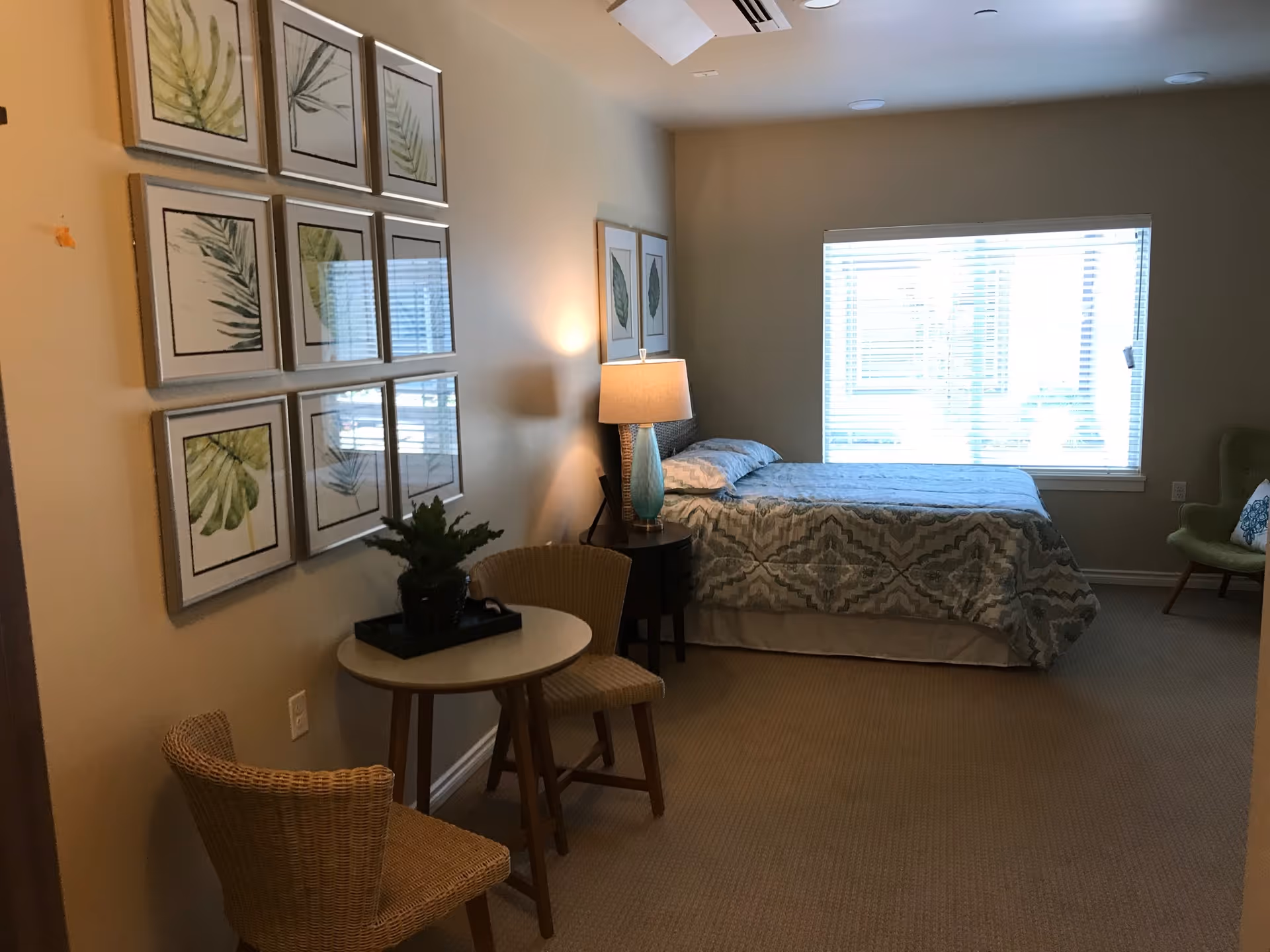 A cozy bedroom with a bed covered in a patterned comforter positioned near a window with blinds. There is a nightstand with a lamp next to the bed, a small round table with two wicker chairs, and a green armchair with a decorative pillow. The walls are adorned with framed botanical prints.