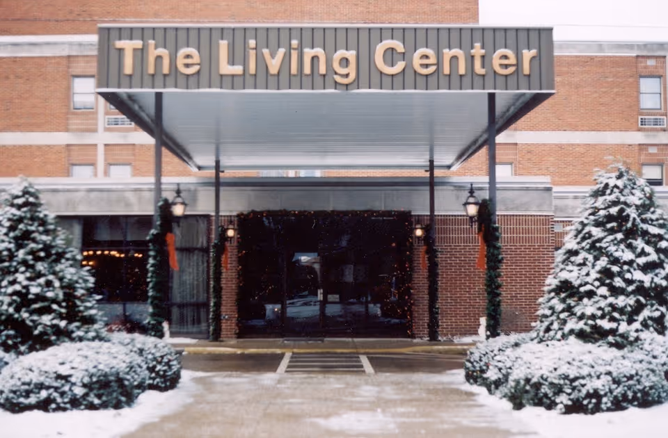 Entrance of The Living Center building with a covered walkway. Snow-covered bushes and trees flank the walkway leading to glass doors. The building is made of red brick with beige accents.