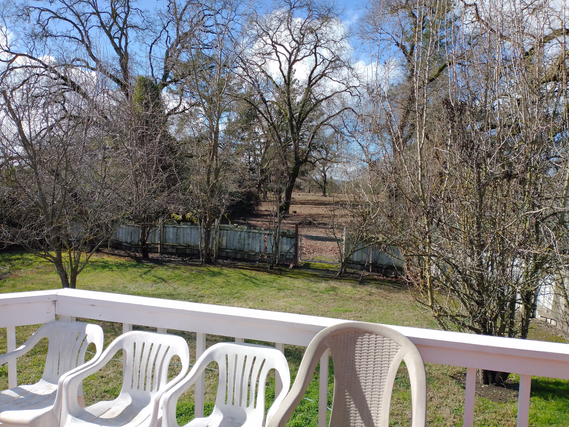 View from a white wooden balcony with four white plastic chairs, overlooking a grassy yard with leafless trees and a weathered wooden fence under a partly cloudy blue sky.