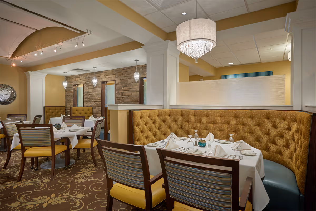 A cozy dining area in a retirement facility with tables set for meals. The room features a curved mustard-colored tufted banquette, wooden chairs with yellow cushions, white tablecloths, and neatly arranged silverware and glassware. The walls are painted a warm beige with stone accent walls and decorative hanging lights. A chandelier hangs from the ceiling, adding an elegant touch.