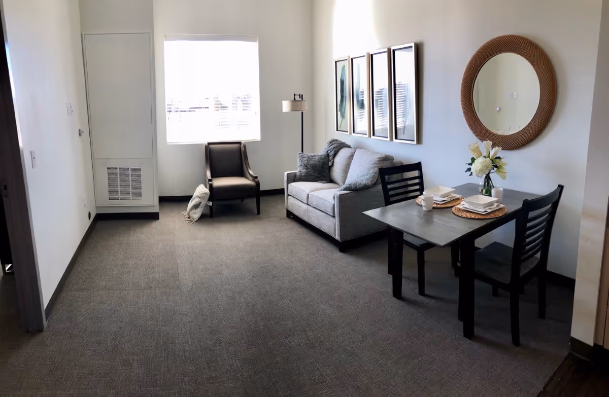 A small living area with a gray loveseat, a dark armchair, a floor lamp, and a small dining table set for two with white dishes and a vase of flowers. There are four framed pictures on the wall above the loveseat and a round mirror on the adjacent wall. The room has a window with blinds letting in natural light.