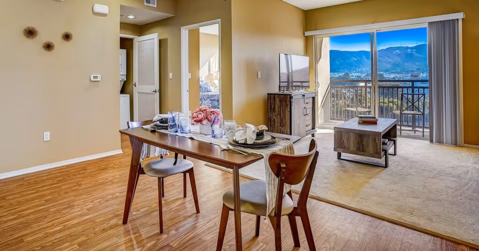 Interior view of a senior living facility apartment at Peninsula Del Rey showing a dining area with a wooden table set for two, a living room with a TV on a wooden stand, a coffee table, and a sliding glass door leading to a balcony with a scenic mountain view. A bedroom is visible through an open door, and a stacked washer and dryer are partially visible behind a louvered door.
