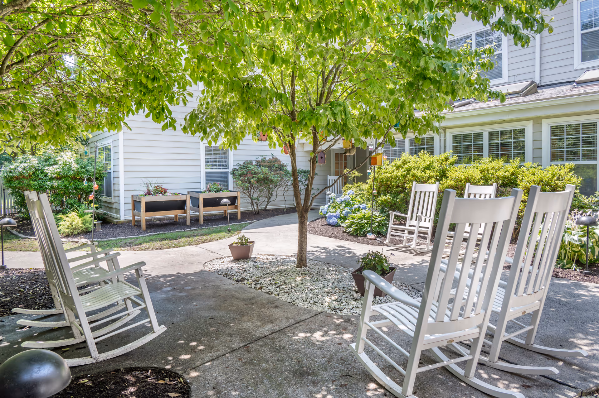 A peaceful outdoor garden area at Brookdale Wilton featuring white wooden rocking chairs arranged around a tree with green leaves. The garden includes flower beds, potted plants, and a paved walkway. The building exterior with white siding and multiple windows is visible in the background.