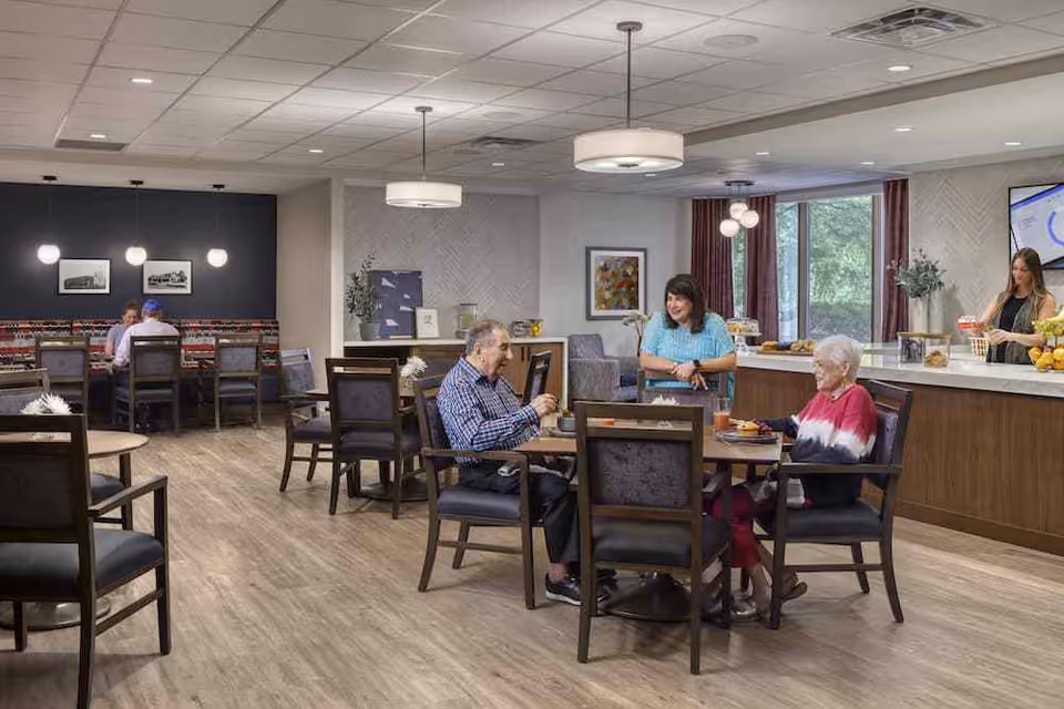 A bright and spacious dining area in Knollwood Place Apartments with several elderly people seated at tables enjoying a meal and conversation. The room features wooden flooring, modern pendant lights, and large windows with curtains allowing natural light. A woman stands behind a counter with various food items, and there are framed pictures on the walls.