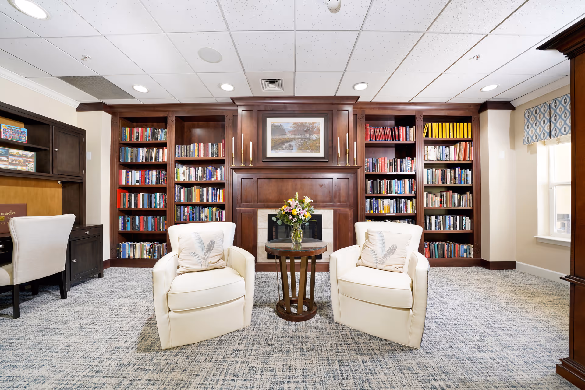 A cozy library room with two white armchairs facing a small round wooden table with a vase of flowers. Behind the chairs is a wooden fireplace mantel flanked by built-in bookshelves filled with books. To the left, there is a desk with a chair, and to the right, a window with patterned valance curtains. The room has a carpeted floor and a white drop ceiling with recessed lighting.