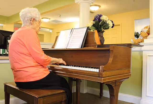An elderly woman with white hair and glasses wearing a coral long-sleeve shirt and black pants is seated on a wooden bench playing a brown upright piano in a well-lit room with light green walls. There is sheet music on the piano and a vase with purple and white flowers on top. A teddy bear is visible on a ledge in the background.