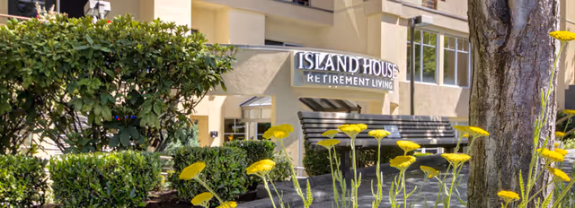 Exterior view of Island House Retirement Living building with a garden area featuring green bushes, yellow flowers, a tree trunk, and a bench in the foreground.