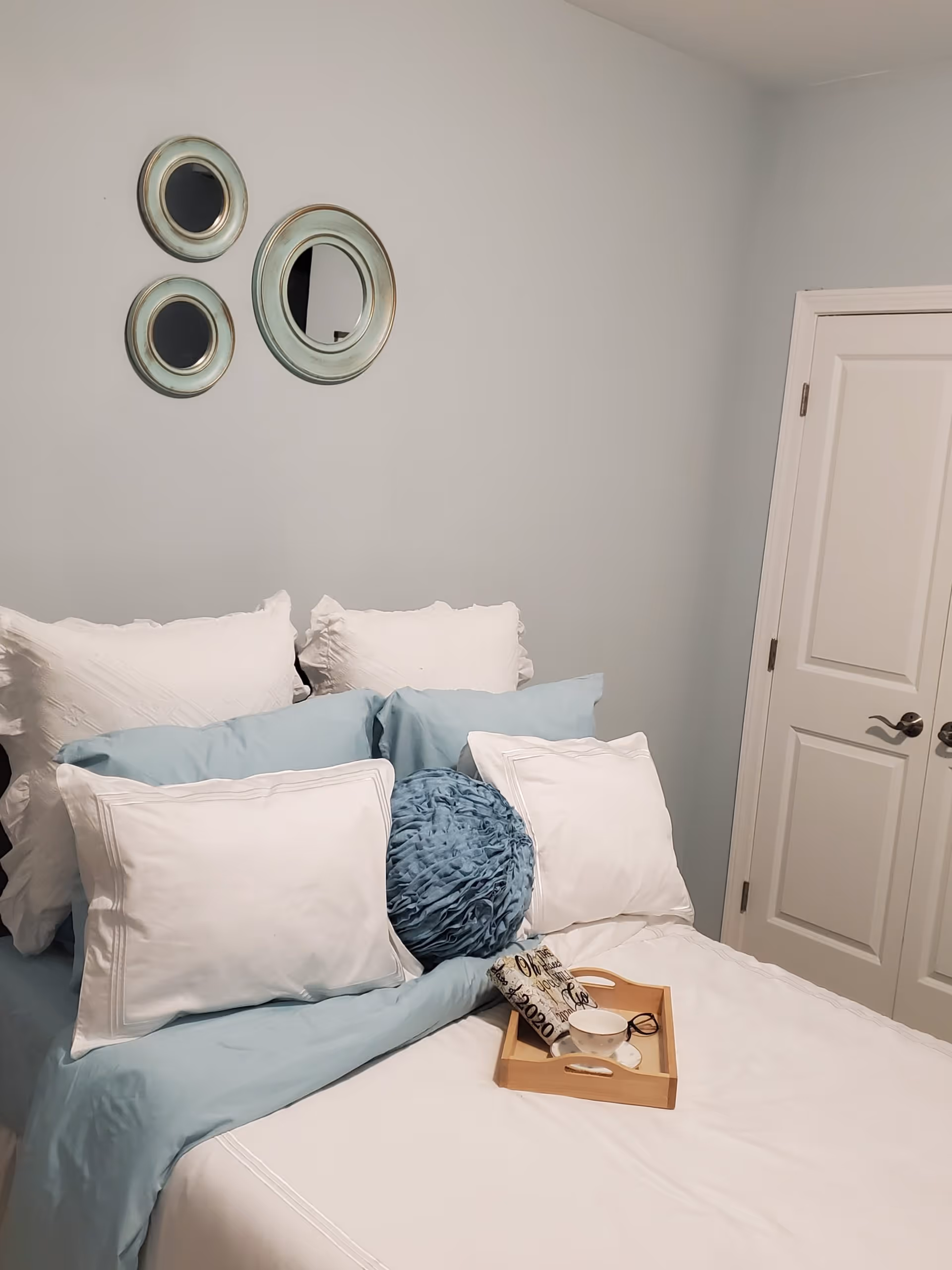 A neatly made bed with white and light blue bedding, including multiple pillows and a round blue decorative pillow. On the bed is a wooden tray holding a cup, a pair of glasses, and a small decorative item. The wall behind the bed is light blue with three round mirrors arranged in a triangular pattern. A closed white door with silver handles is visible on the right side.