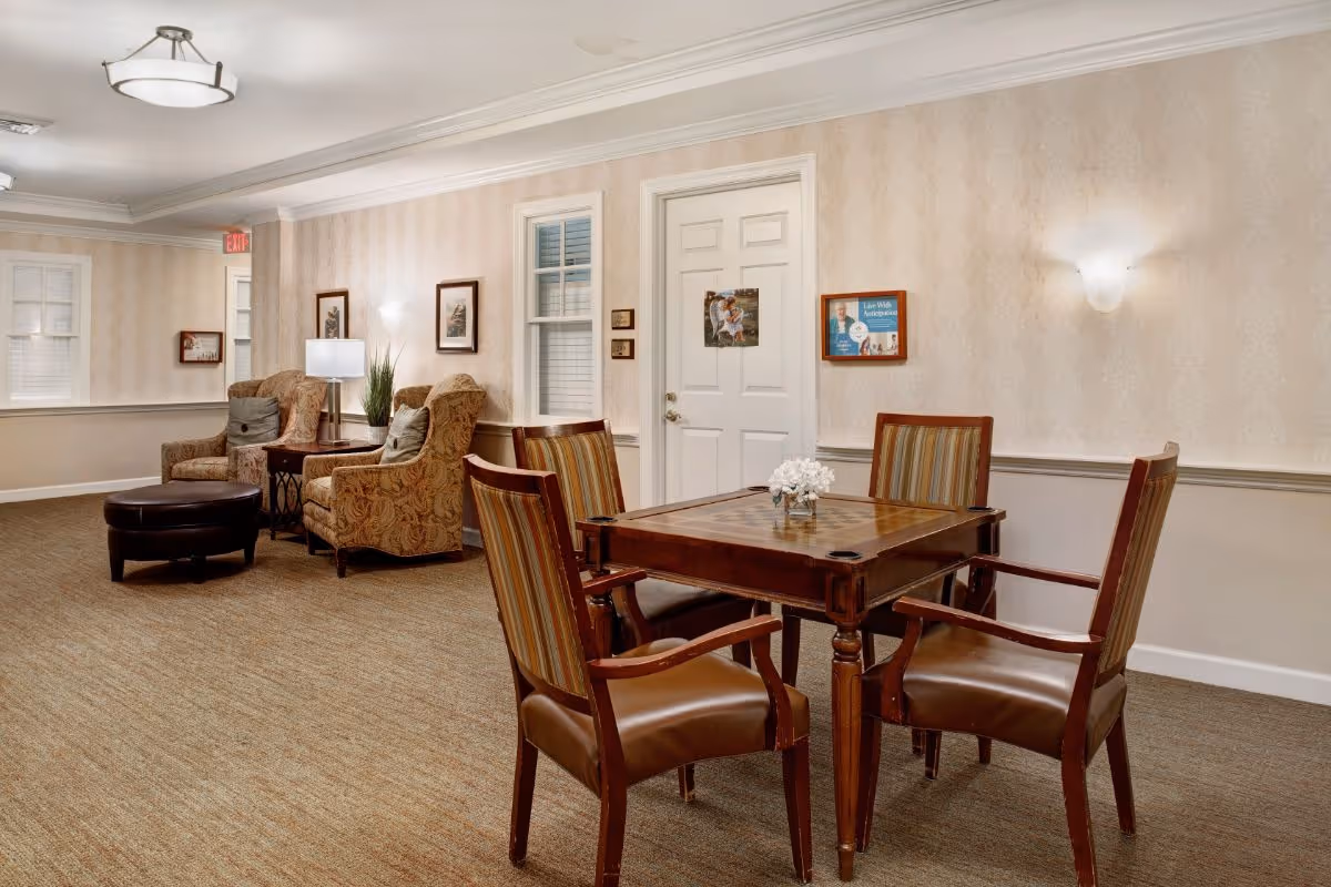 A warm common room with a wooden game table and four chairs, upholstered armchairs, ottoman, and soft wall lighting.