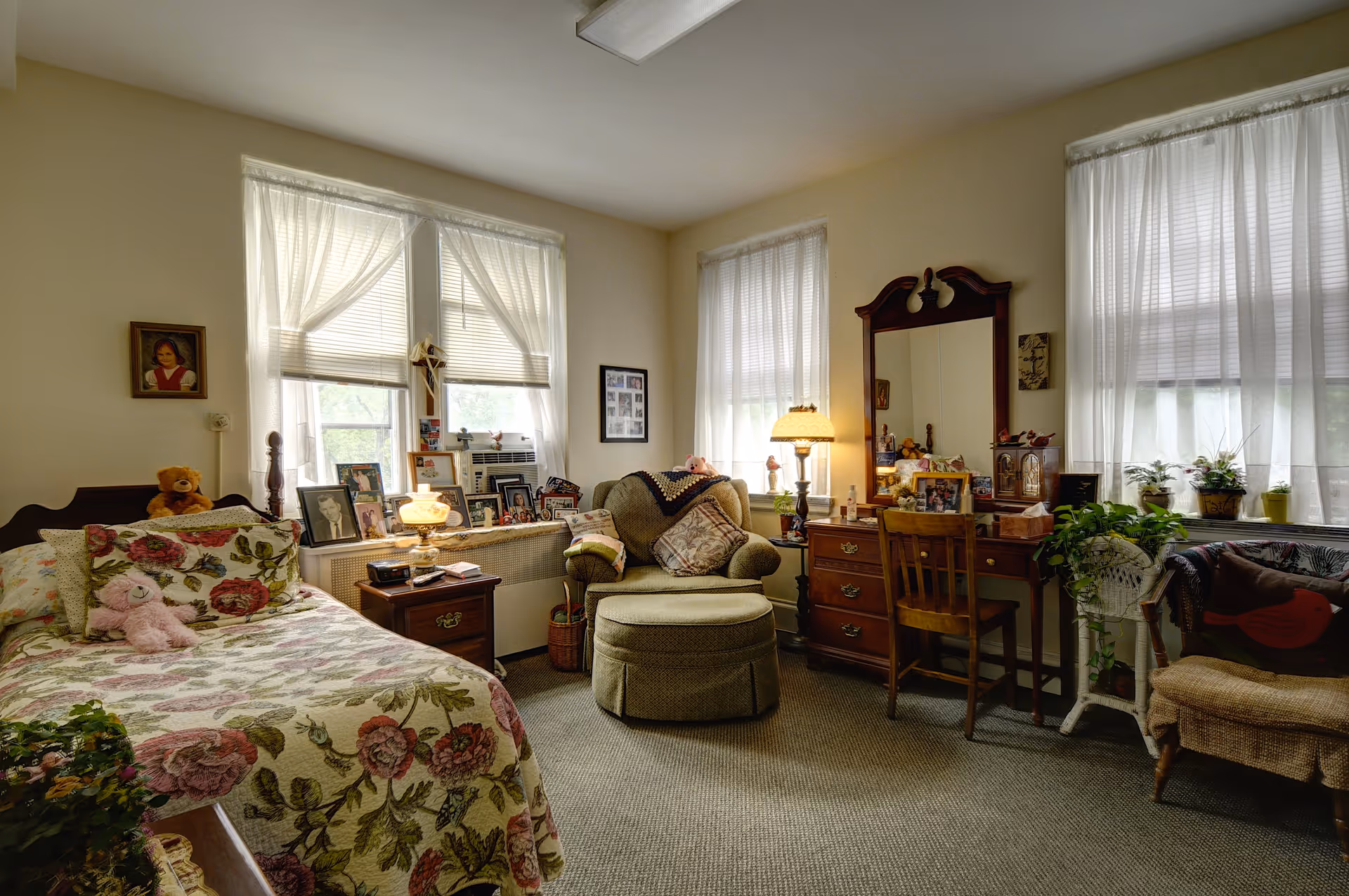 A cozy bedroom in Saint Francis Residential Community featuring a bed with floral bedding and stuffed animals, a cushioned armchair with an ottoman, a wooden dresser with a mirror, a small desk with a chair, several framed photos on the windowsill, and sheer curtains covering three windows letting in natural light.