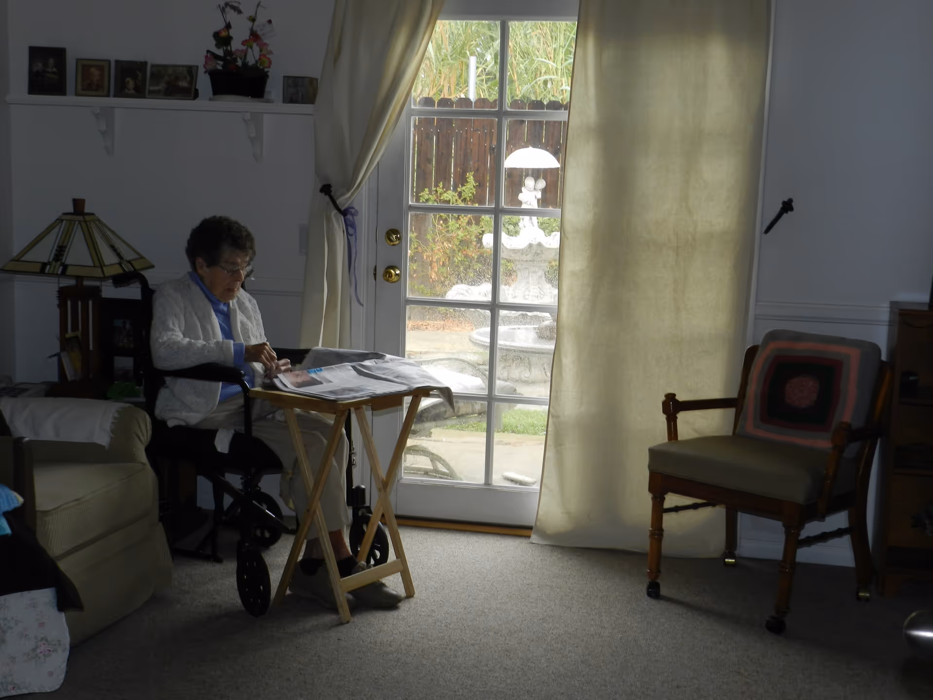 An elderly woman sitting in a wheelchair inside a living room, reading a newspaper on a small wooden tray table. The room has a glass door with curtains partially drawn, showing a garden with a decorative fountain outside. There is a cushioned wooden chair with a crocheted pillow and a lamp on a side table nearby.