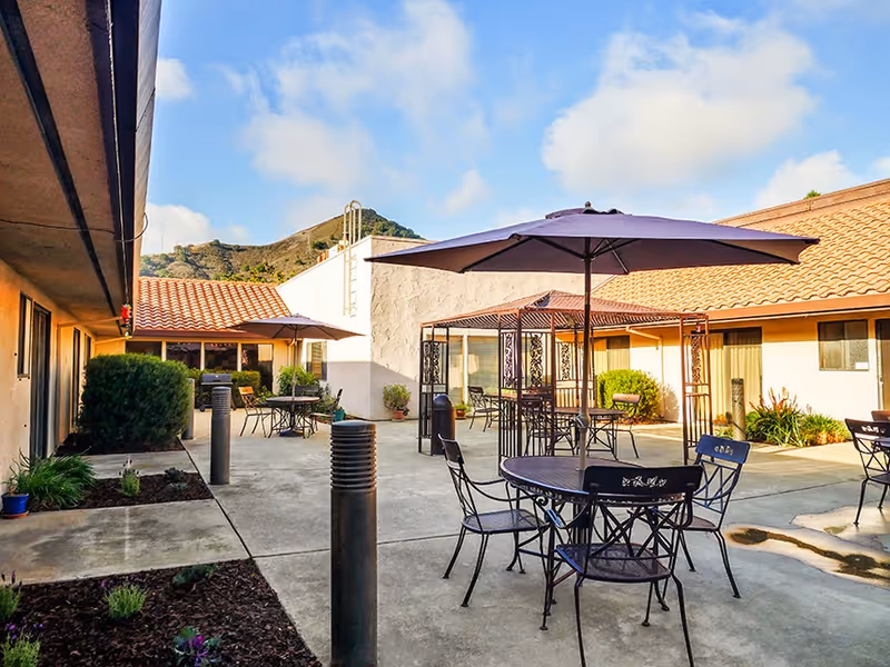 Sunny courtyard with metal tables, chairs, umbrellas and a gazebo between single-story buildings with tiled roofs.