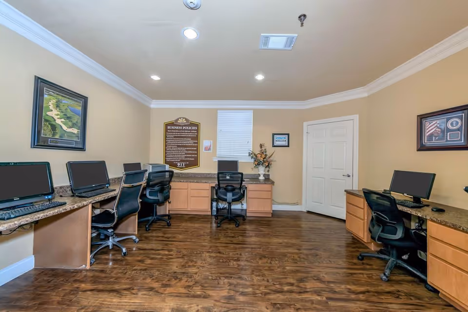 A computer room with multiple workstations featuring desktop computers and office chairs arranged along the walls. The room has wooden flooring, beige walls, and framed pictures and a business policies sign on the walls. There is a window with blinds in the center of the back wall and a closed white door to the right.