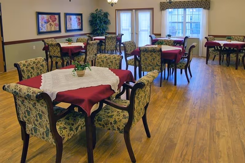 A dining room with several tables covered with red tablecloths and white lace overlays, surrounded by floral upholstered chairs. The room has wooden flooring, framed floral artwork on the walls, a large potted plant, and windows with sheer curtains allowing natural light to enter.