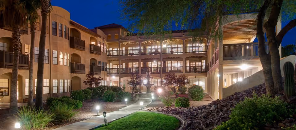 Courtyard and front facade of the Amber Lights senior living building at dusk, with illuminated balconies, a landscaped walkway, and a central fountain.