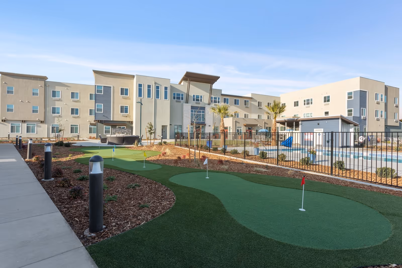 Outdoor view of WellQuest of Elk Grove facility showing a putting green with multiple small flags, a fenced swimming pool area with a slide, palm trees, and a modern three-story building in the background under a clear blue sky.
