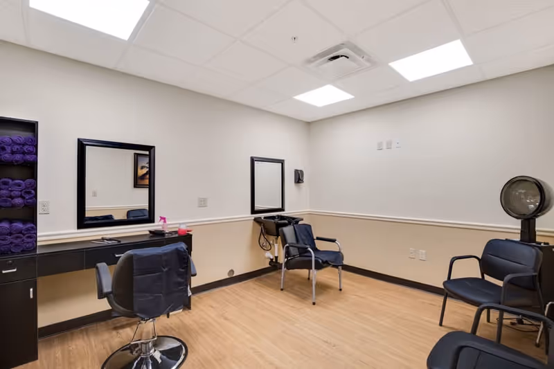 Interior view of a salon area in an assisted living facility with a styling chair in front of a counter with two mirrors, a black sink for hair washing, several black chairs, and shelves stocked with purple towels. The room has light-colored walls and wood flooring with a white ceiling and fluorescent lights.